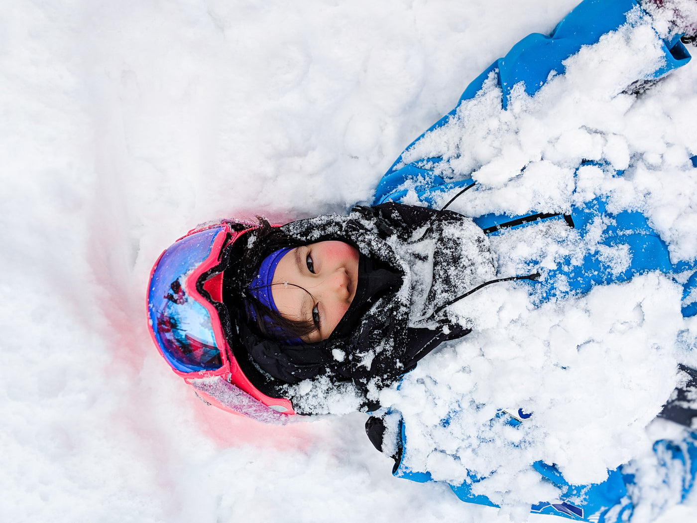 Smiling Girl in Blue Ski Jacket with Pink Helmet and Ski Goggles, Lying Half Buried In Snow