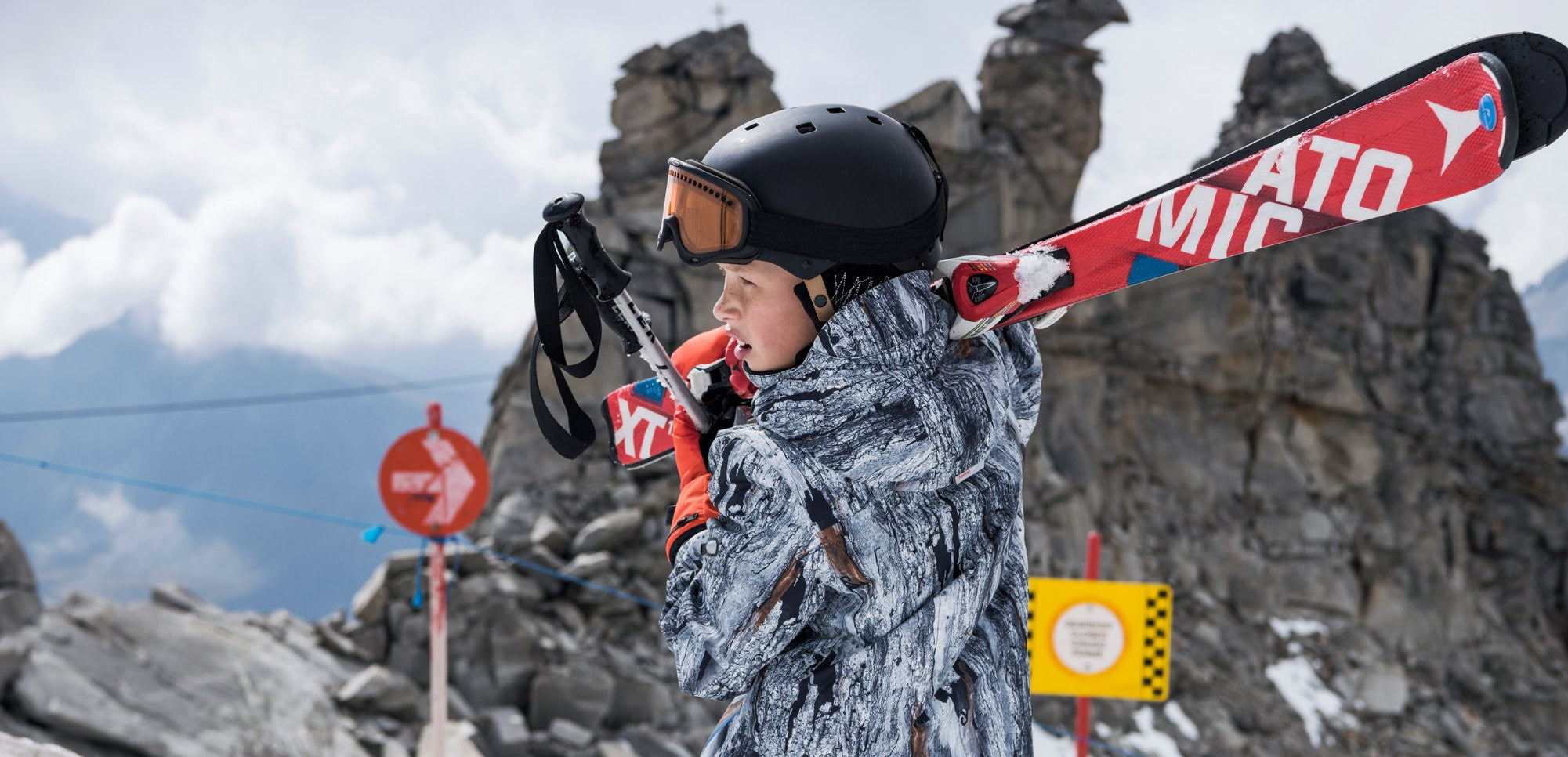 teenager in colourful snow gear wearing a ski helmet and goggles carrying skis