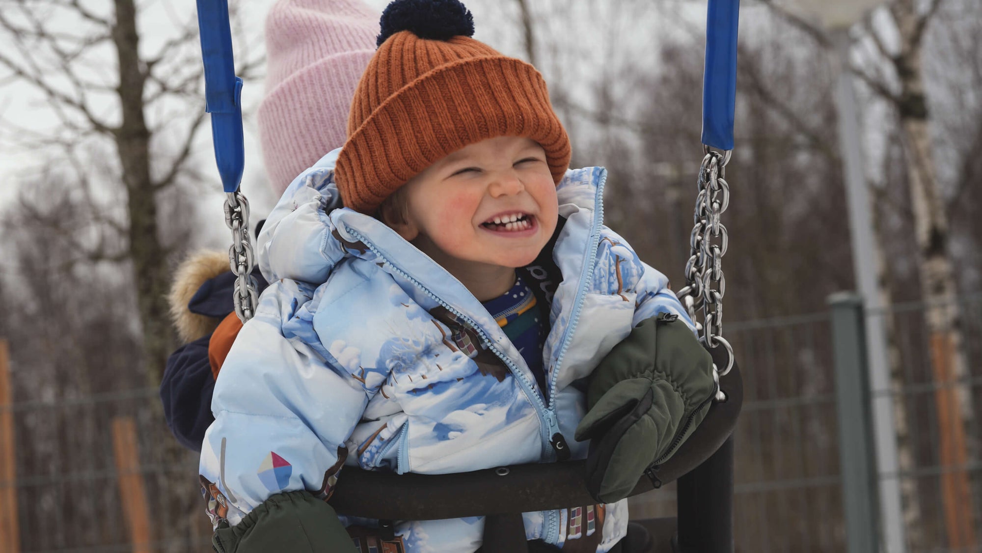 Child in winter clothing being pushed on a swing in a snowy playground.
